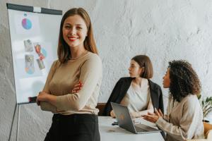 Jeune femme souriante au bureau, avec deux collègues en arrière-plan. Ambiance de travail détendue et collaborative.